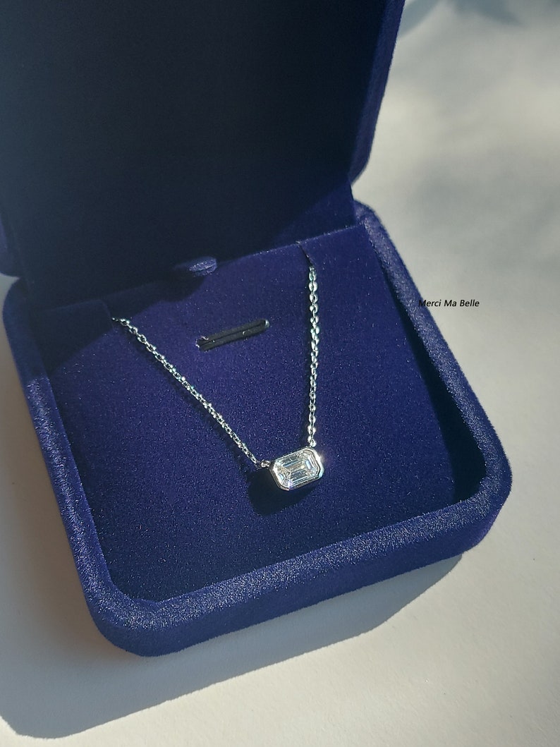 May include: A silver necklace with a rectangular, clear gemstone pendant in a blue velvet box. The box has the text "Merci Ma Belle" printed on it.