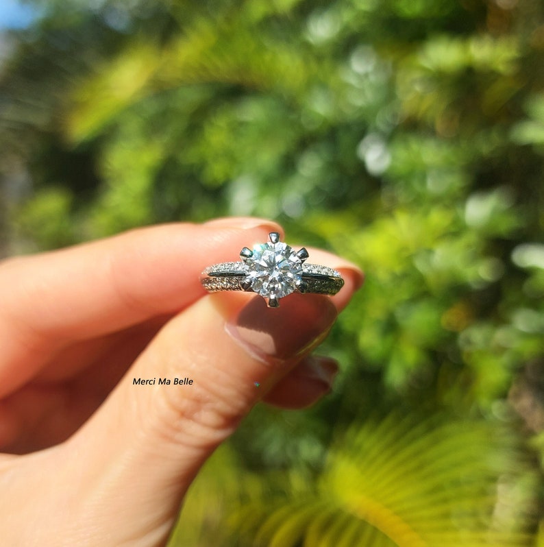 May include: A silver engagement ring with a large, round, clear diamond center stone and smaller diamonds set in the band. The ring is on a finger against a blurred green background.