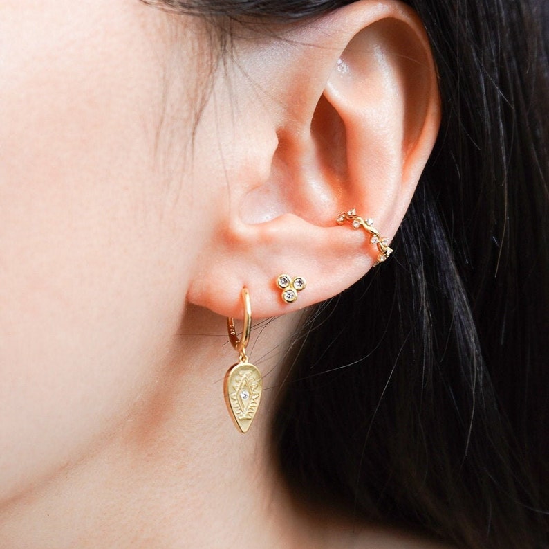 May include: Gold hoop earrings with a teardrop-shaped charm featuring a detailed eye design and a small diamond. The earrings are shown on a person's ear with a small cluster of diamonds and a gold ear cuff.