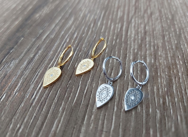May include: Three pairs of earrings with a teardrop-shaped charm featuring an evil eye design. The charms are gold and silver and have a small clear stone in the center of the eye.