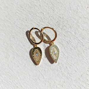 May include: Gold hoop earrings with a teardrop-shaped pendant featuring a geometric design and a small clear stone in the center.