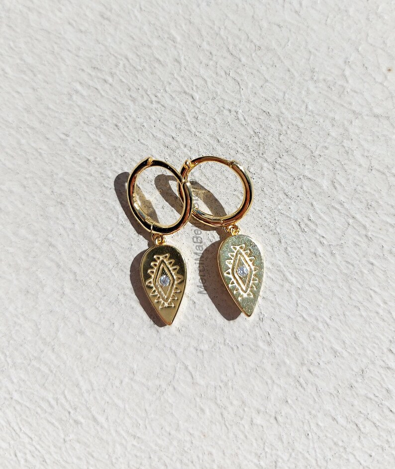 May include: Gold hoop earrings with a teardrop-shaped pendant featuring a geometric design and a small clear stone in the center.