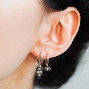 May include: A pair of silver hoop earrings with a small silver charm hanging from each hoop. The charm on the left is a teardrop shape with a diamond-shaped design and a small diamond in the center. The charm on the right is a small eye with a diamond in the center.