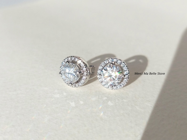 May include: A pair of silver stud earrings with a round, clear stone surrounded by a halo of smaller stones.  Merci Ma Belle Store is printed in the background.