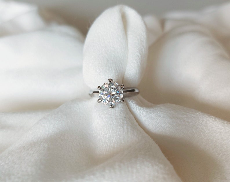 May include: A silver ring with a large, clear, round diamond set in a six-prong setting.