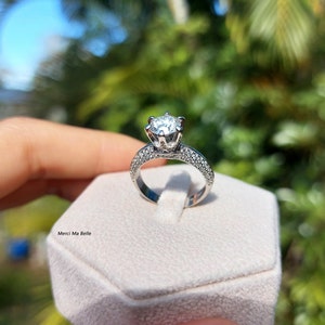 May include: A silver engagement ring with a large, round, clear diamond center stone and smaller diamonds set in a pave setting around the band. The ring is displayed in a white ring box.