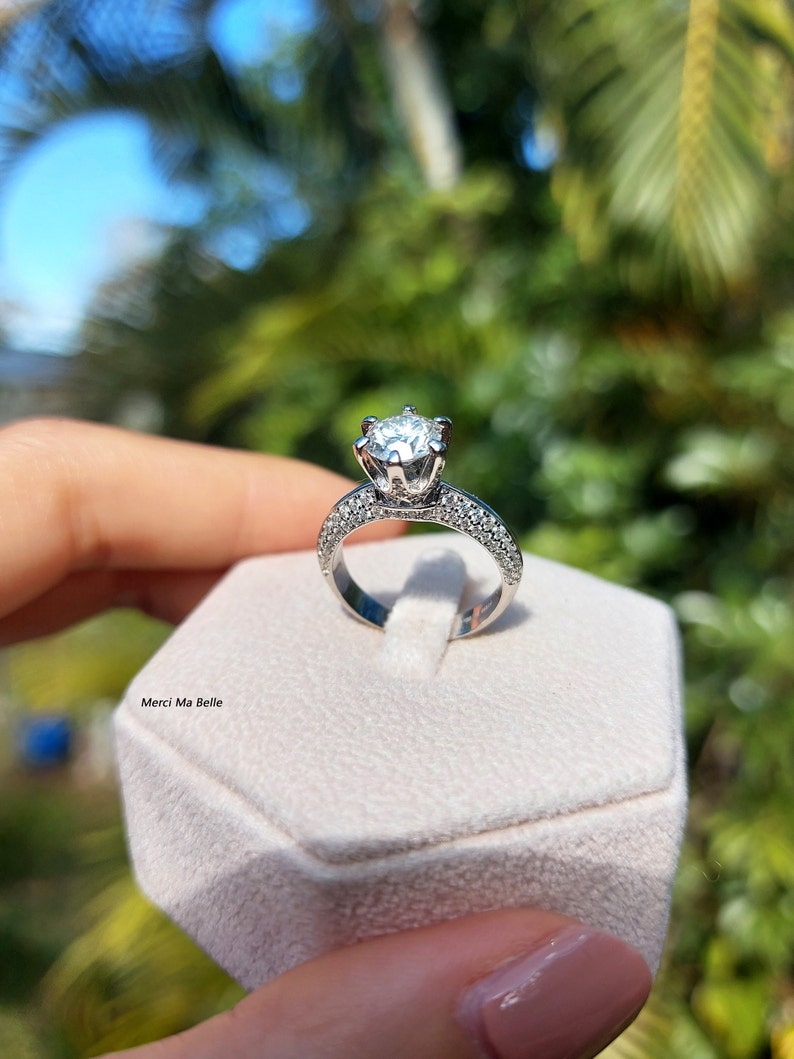 May include: A silver engagement ring with a large, round, clear diamond center stone and smaller diamonds set in a pave setting around the band. The ring is displayed in a white ring box.