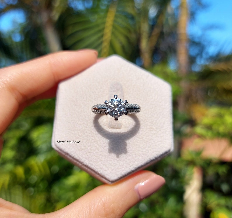 May include: A silver ring with a large, round, clear diamond in the center. The ring has a double row of smaller diamonds around the center stone. The ring is in a white ring box with the words "Merci Ma Belle" printed on the bottom.