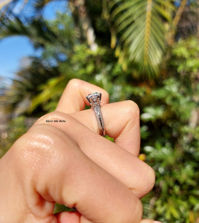 May include: A silver engagement ring with a large, round, clear diamond center stone. The band is set with smaller diamonds and has a delicate, intricate design.