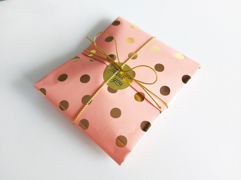 May include: A pink gift bag with gold polka dots and a gold ribbon. A gold sticker with the text "Thank you" is attached to the ribbon.