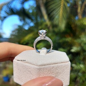 May include: A silver ring with a large, round, clear center stone and smaller stones around the band. The ring is in a white ring box with the text "Merci Ma Belle" on the front.