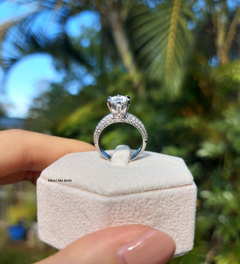 May include: A silver ring with a large, round, clear center stone and smaller stones around the band. The ring is in a white ring box with the text "Merci Ma Belle" on the front.
