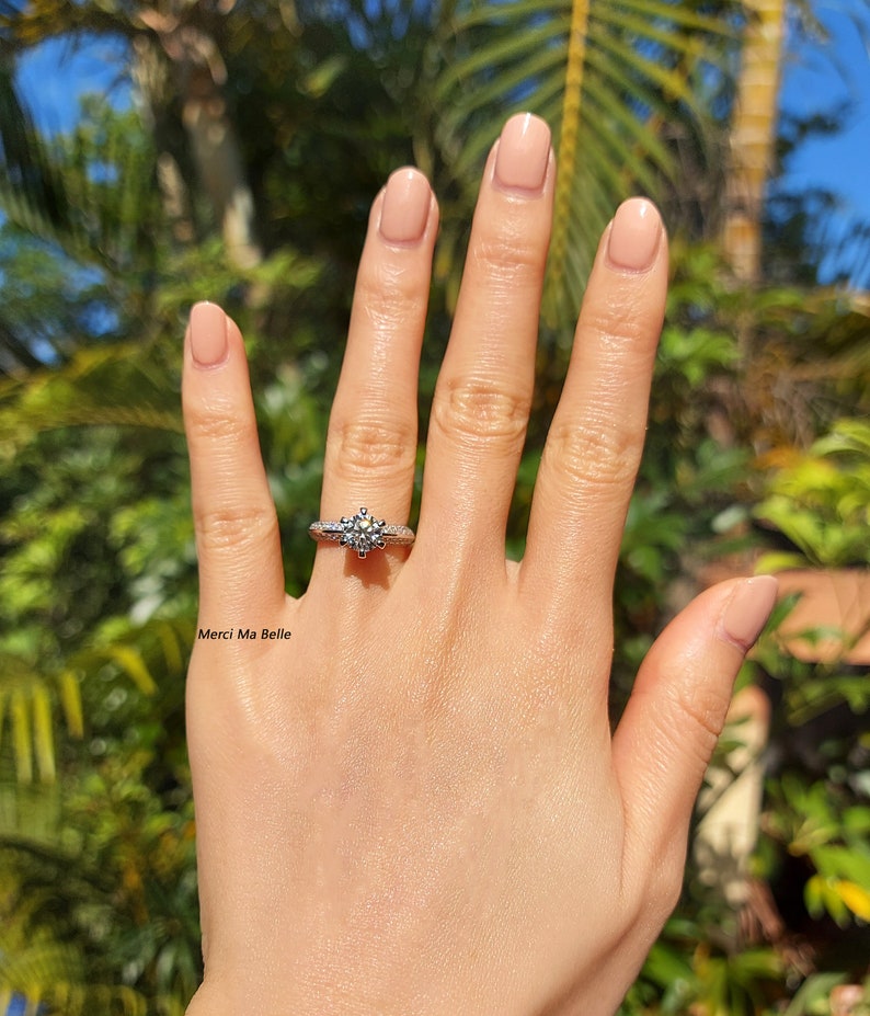 May include: A close-up of a hand wearing a silver ring with a large, round, clear diamond. The ring has a delicate, intricate design. The text "Merci Ma Belle" is visible in the image.