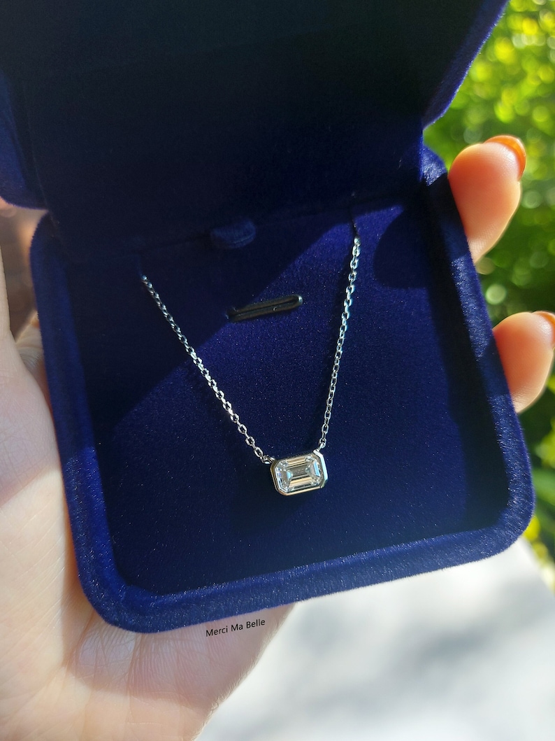 May include: A silver chain necklace with a small, clear, emerald-cut gemstone pendant. The necklace is in a blue velvet box with the words "Merci Ma Belle" printed on the bottom.