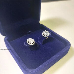 May include: A pair of silver earrings with a round, clear stone surrounded by smaller stones in a halo setting. The earrings are displayed in a blue velvet jewelry box.