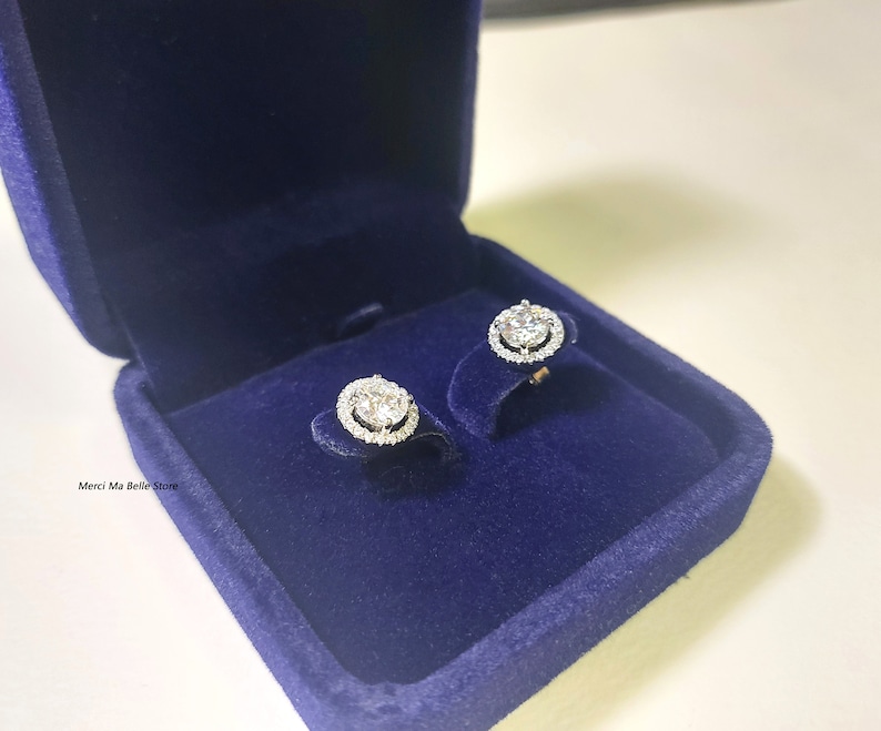 May include: A pair of silver earrings with a round, clear stone surrounded by smaller stones in a halo setting. The earrings are displayed in a blue velvet jewelry box.