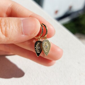May include: Gold hoop earrings with a dangling teardrop-shaped charm featuring a geometric design and a small clear rhinestone.
