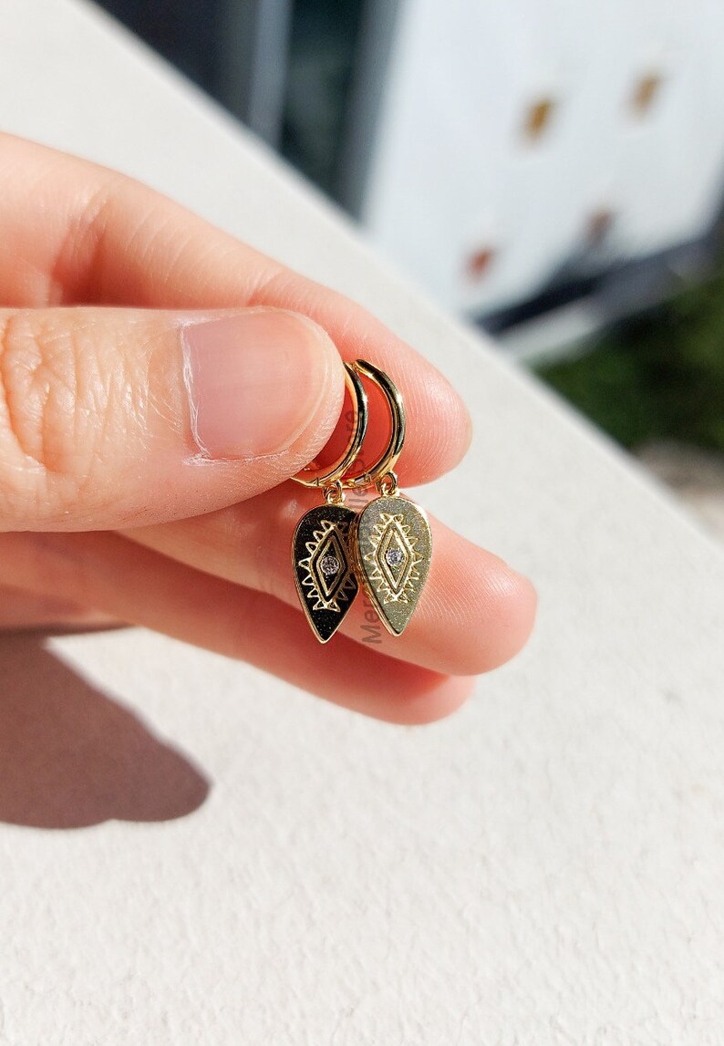 May include: Gold hoop earrings with a dangling teardrop-shaped charm featuring a geometric design and a small clear rhinestone.