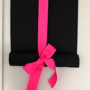 May include: A rectangular black fabric wall hanging suspended from a wooden dowel. A wide, vibrant neon pink ribbon runs vertically down the center of the black fabric, tied into a decorative bow at the bottom where the fabric is rolled upward.