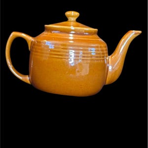 May include: A ceramic teapot in a warm, amber-brown colour. The teapot features a rounded body with horizontal ridges, a curved handle, and a spout. The lid has a small knob. The background is black.