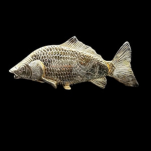 Puede incluir: Una detallada escultura de pez metálico con escamas y aletas texturizadas. La escultura es de color dorado y está sobre un fondo negro. El pez está orientado horizontalmente, mostrando su intrincado diseño.