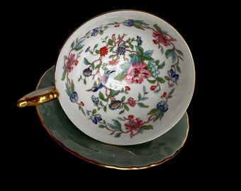 May include: White teacup and saucer set with a floral and bird design. The teacup has a gold handle and rim, and the saucer is a muted green with a gold trim. The floral pattern includes pink, red, blue, and green elements.