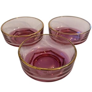 Peut inclure: Trois bols en verre transparent avec un bord doré et une base magenta foncé. Les bols ont un motif géométrique au centre. Les bols sont probablement destinés à servir des desserts ou des apéritifs.