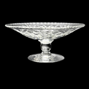 Puede incluir: Una fuente de vidrio transparente con pie, con un cuenco ancho y poco profundo y un patrón decorativo de vidrio tallado. La fuente tiene una base redonda y un tallo corto y redondeado. El borde del cuenco tiene un borde festoneado.