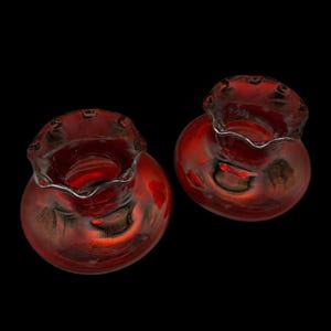 Può includere: Due piccoli vasi rotondi in vetro rosso con bordi arricciati e finiture argentate. I vasi hanno un corpo bulboso e un'ampia apertura. Il vetro ha una finitura lucida e riflette la luce.