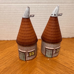 May include: Two ceramic house-shaped ornaments. Each has a brown, textured roof, a grey cylindrical base with a green door and yellow window. The chimney is white with a small, curved extension.