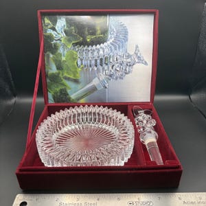 May include: A clear glass ashtray with a radial pattern and a matching glass bottle stopper with a grape cluster design, presented in a burgundy velvet-lined box. A stainless steel ruler shows the set measures approximately 23 cm in width.