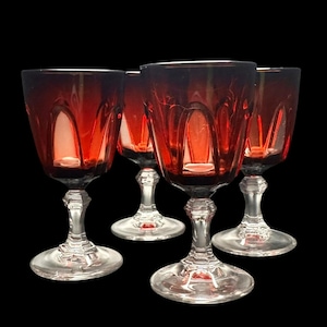 May include: Four red wine glasses with clear stems and bases. The glasses have a decorative pattern on the bowl and a classic design. The glasses are set against a black background.