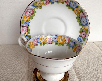 Puede incluir: Juego de taza y platillo blancos con diseño floral en amarillo, rosa, azul y verde. La taza está sobre un soporte dorado. El borde de la taza y el platillo tiene un ribete dorado.