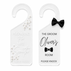 May include: Two clear acrylic door hangers. The left hanger features white text reading PREPARING to be MRS Smith, PLEASE KNOCK, and is decorated with white pearls. The right hanger features black text reading THE GROOM Oliver's ROOM, PLEASE KNOCK, and a black bow tie.