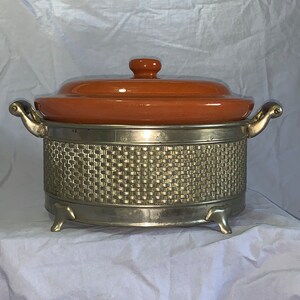 May include: A vintage orange ceramic casserole dish with a silver metal base and woven design. The dish has two handles and sits on three feet.