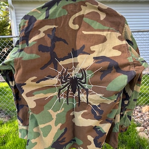 May include: A camouflage green, brown, and black utility jacket with a white spider web and black spider embroidered on the back.