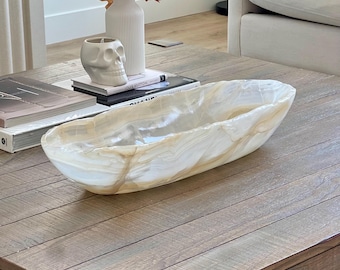 May include: A large, oval-shaped decorative bowl with a polished, marbled appearance. The bowl is primarily white with streaks of tan and gold. It sits on a wooden coffee table, alongside books, a skull-shaped candle holder, and a white vase with fall foliage.