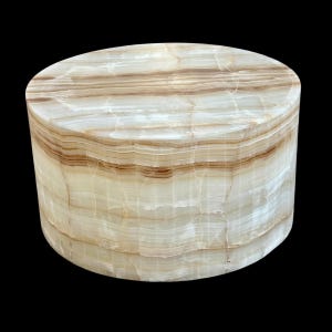 May include: A cylindrical decorative object made of layered, translucent onyx. The natural stone features bands of cream and brown hues, creating a striped pattern. The object has a smooth, polished surface and a rounded top.
