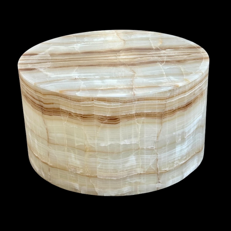May include: A cylindrical decorative object made of layered, translucent onyx. The natural stone features bands of cream and brown hues, creating a striped pattern. The object has a smooth, polished surface and a rounded top.