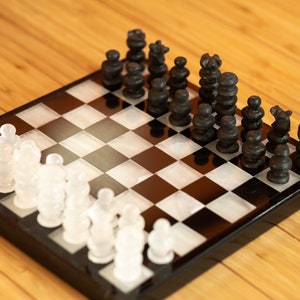 Könnte beinhalten: Ein Schachspiel mit schwarzen und weißen Figuren auf einem Schachbrett. Die schwarzen Figuren sind dunkel, während die weißen Figuren durchscheinend sind. Das Brett steht auf einer Holzoberfläche.