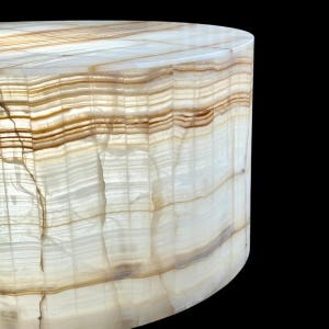 May include: A cylindrical, translucent onyx side table. The table features horizontal bands of cream, tan, and brown, creating a layered effect. The surface is smooth and polished, with a natural, organic aesthetic. The background is black.