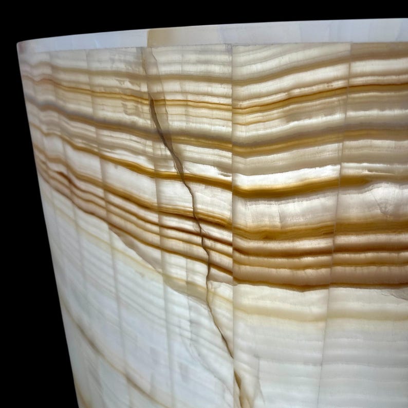 May include: Close-up of a translucent, cylindrical object with a layered pattern. The object features bands of white and brown hues, creating a natural, textured appearance. The material appears to be a type of stone or mineral, possibly onyx.