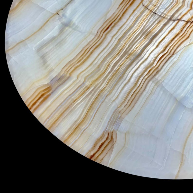 May include: Close-up of a round, polished stone surface with a natural pattern. The surface features a blend of white, cream, and brown tones, with linear striations creating a layered effect. The stone has a smooth, glossy finish.
