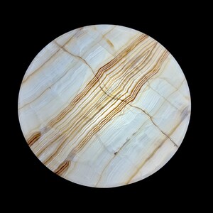 May include: A round, polished stone with a white and tan striped pattern. The stone has a natural, marbled appearance with brown and tan veining. The circular shape is set against a black background, highlighting the stone's unique design.