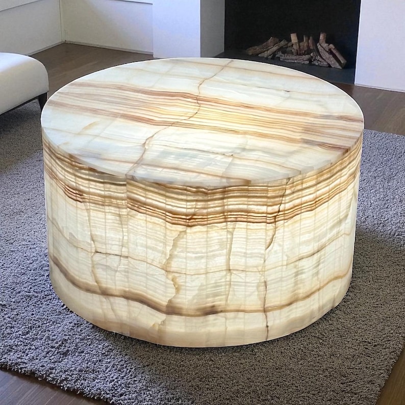 May include: A round, cylindrical coffee table made of a translucent, light-colored stone. The table has a banded pattern of light and dark brown hues. The table is sitting on a gray rug.
