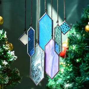 May include: A set of six stained glass ornaments in various shades of blue, purple, and white, hanging from black cords. The ornaments are shaped like elongated hexagons and have a geometric design.
