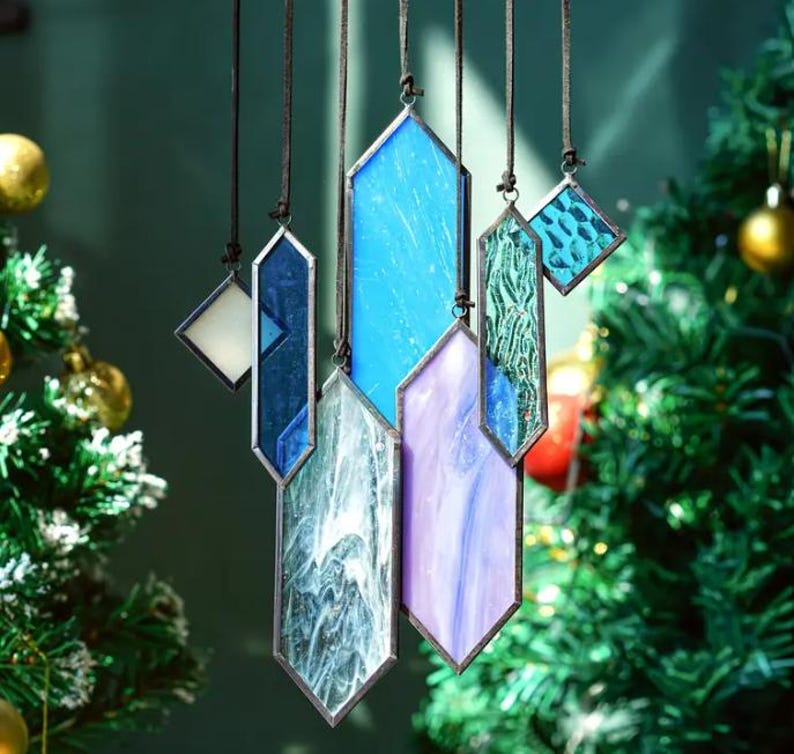 May include: A set of six stained glass ornaments in various shades of blue, purple, and white, hanging from black cords. The ornaments are shaped like elongated hexagons and have a geometric design.