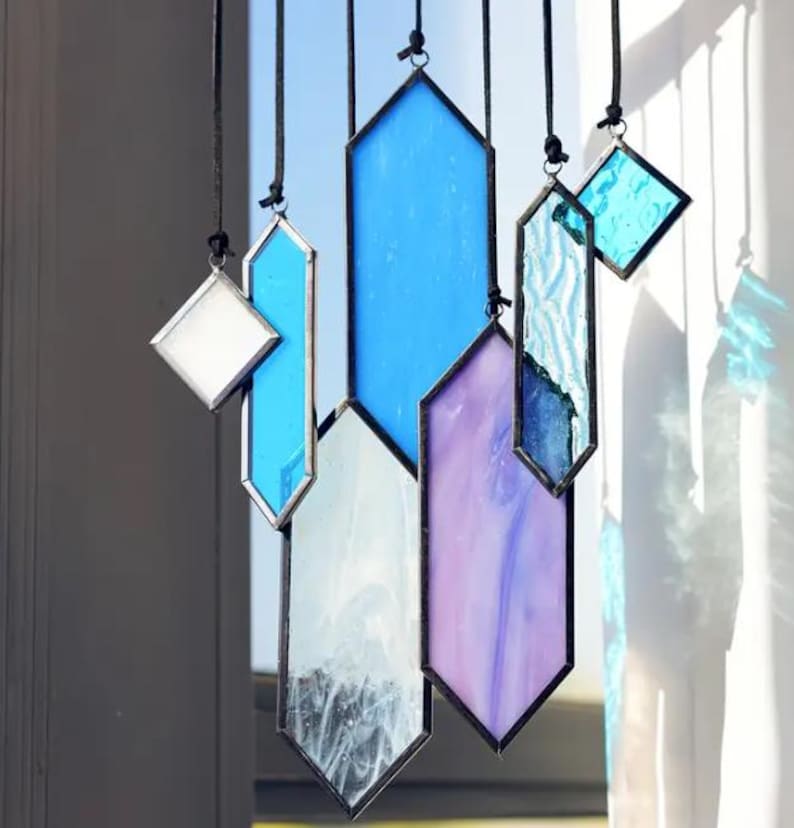 May include: A set of six stained glass window hangings in various shapes and colors, including blue, purple, and white. The hangings are suspended from black cords.