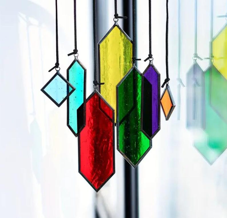 May include: A set of seven stained glass suncatchers hanging from black cords. The suncatchers are in various shapes and colors, including red, green, yellow, blue, purple, and orange.