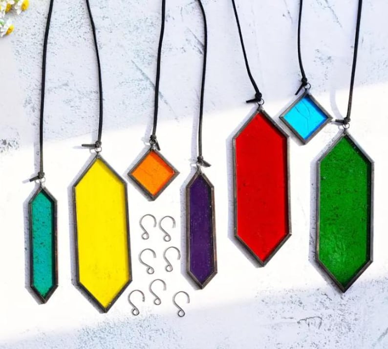 May include: A set of seven stained glass suncatchers, each with a different color and shape. The suncatchers are suspended from black cords and have metal hooks. The colors include green, yellow, orange, red, purple, and blue.